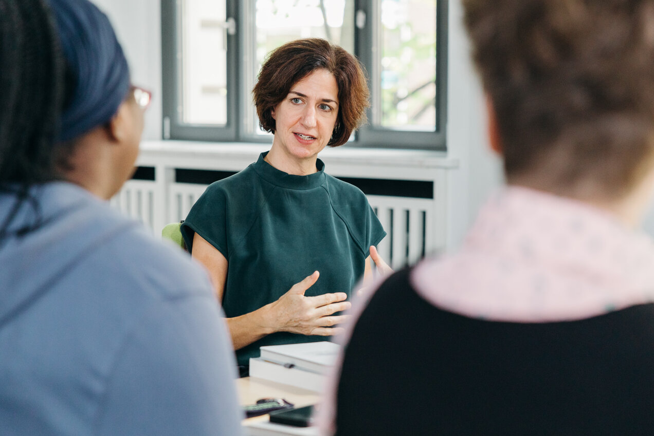 person speaking and gesturing, two persons partly visible in the foreground