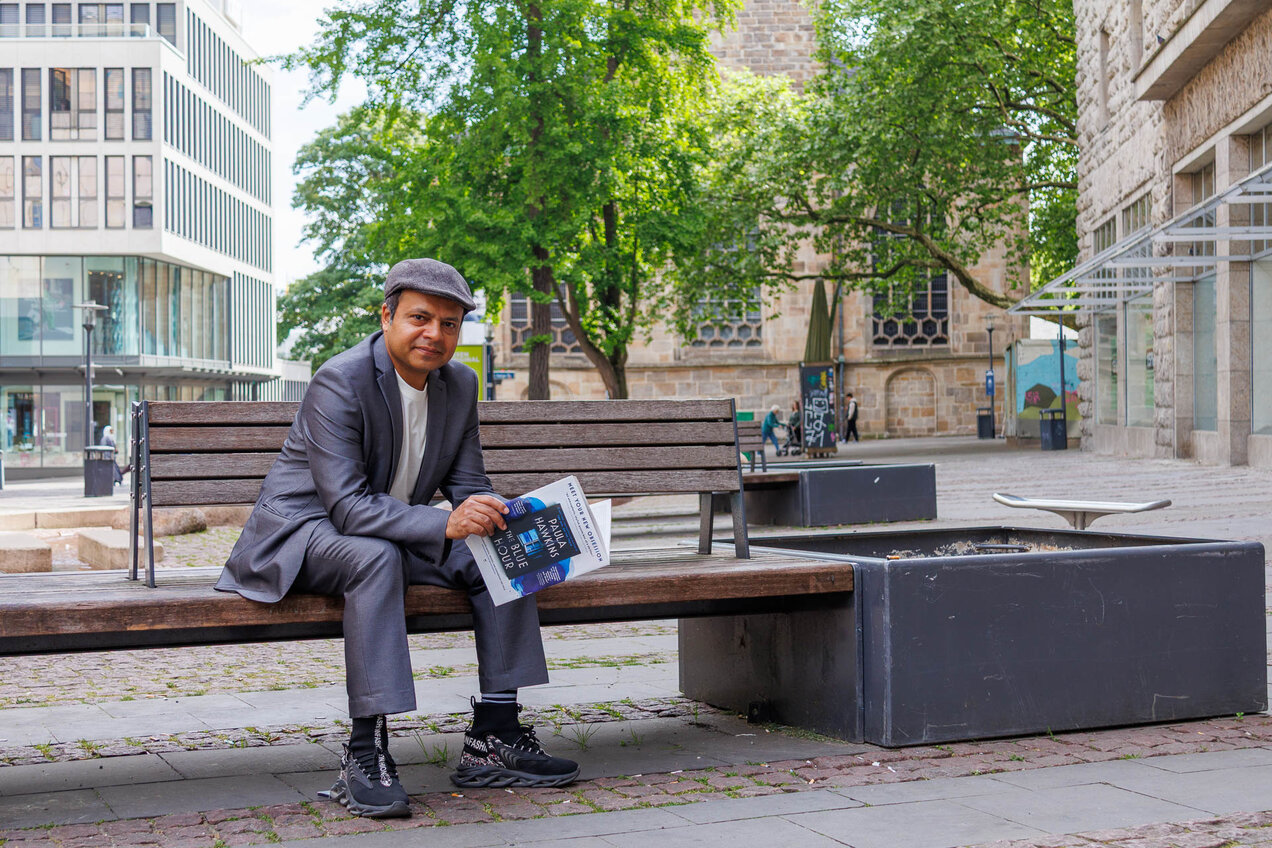 Imran Sabir sitting on a public bench in the city centre of Essen, looking at the camera, holding a newspaper in his hands. In the background there are buildings and trees.