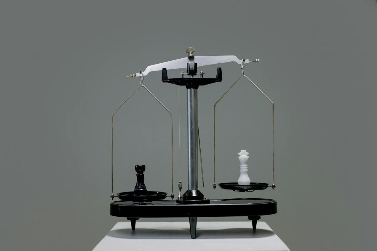 scales with two weighing pans that are not in balance, with a black chess piece on the left side, a white chess piece on the right side