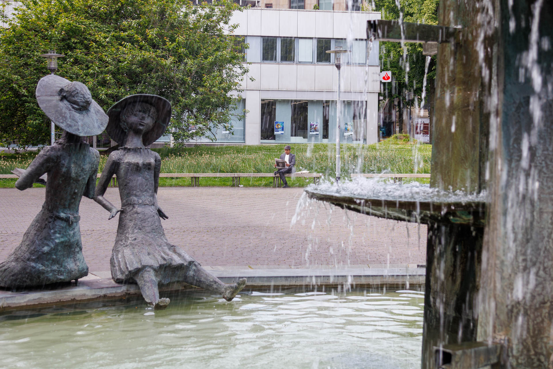 Picture of a fountain with a sculpture of two people sitting on the edge in the foreground. Imran Sabir is sitting on a bench in the background.