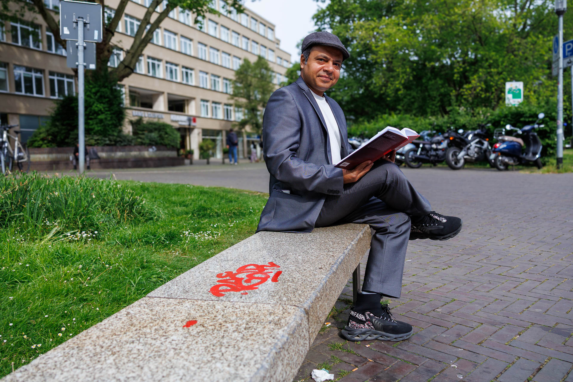 Imran Sabir sitting on a stone bench, holding a book but smiling at the camera. Next to Imran on the bench is red graffiti.