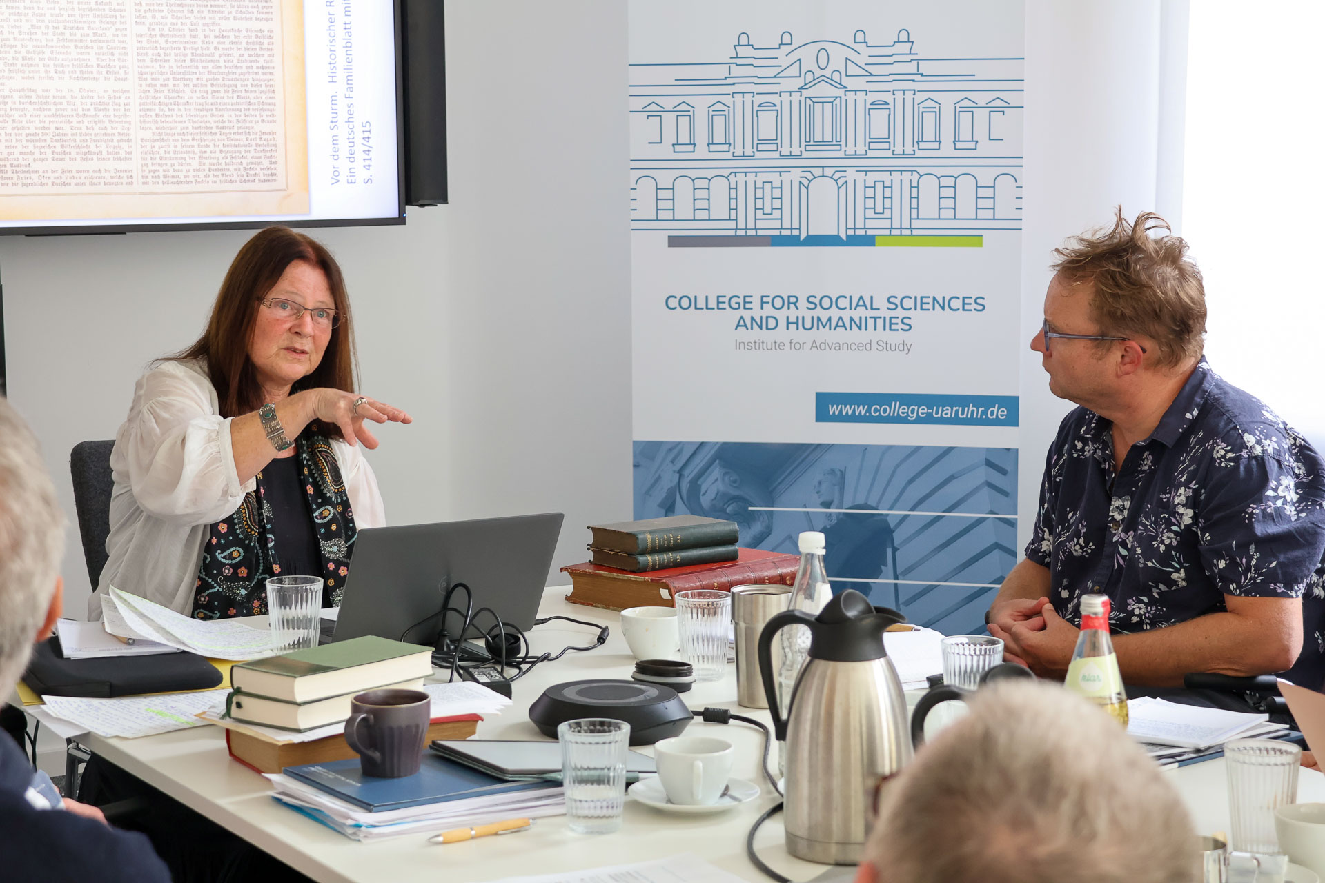 photo from the workshop 'Editing Journal Literature', two participants sitting at a table, a speaker presenting, a screen with slides in the back
