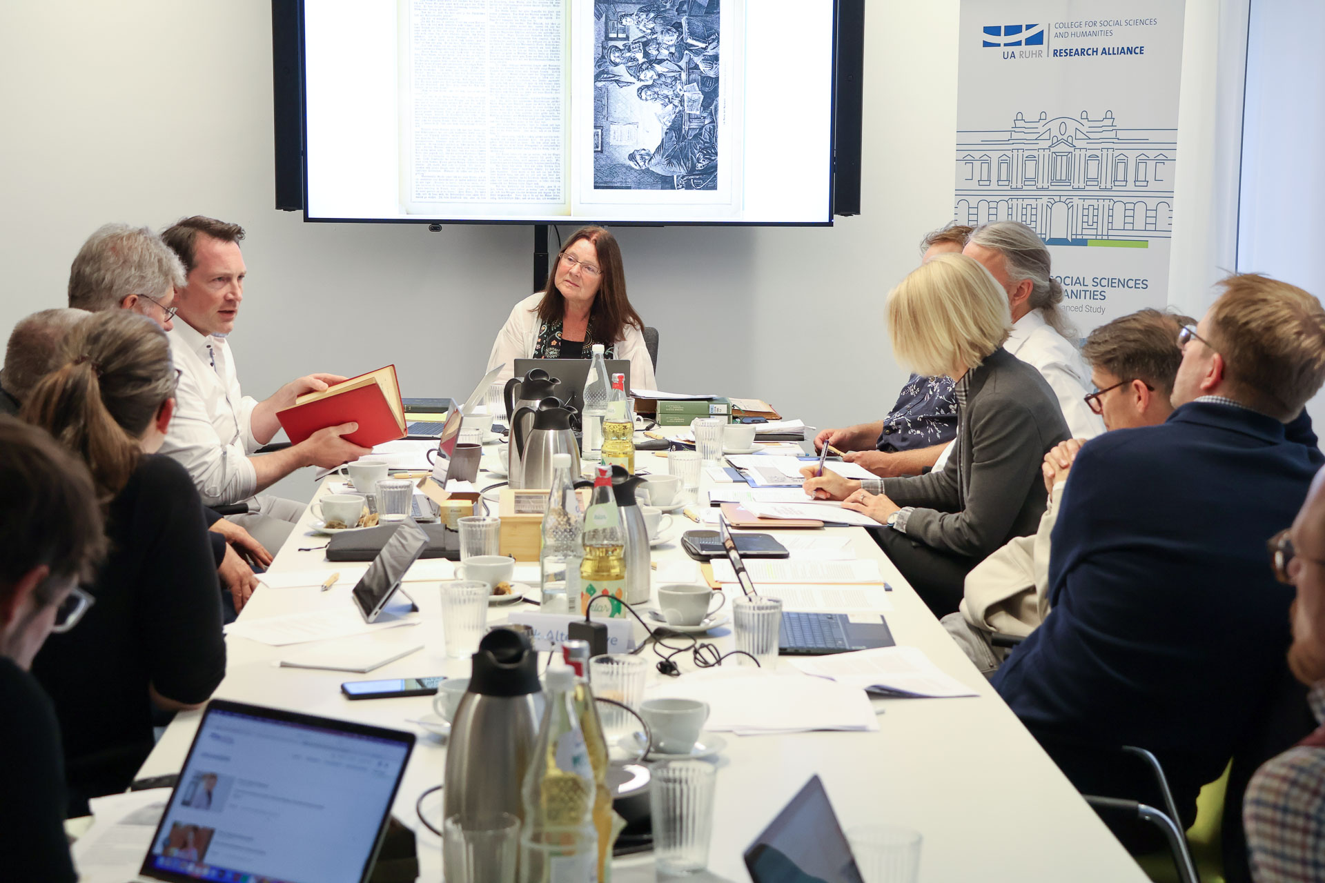 photo from the workshop 'Editing Journal Literature', participants sitting around a table, a speaker presenting, a screen with slides in the back