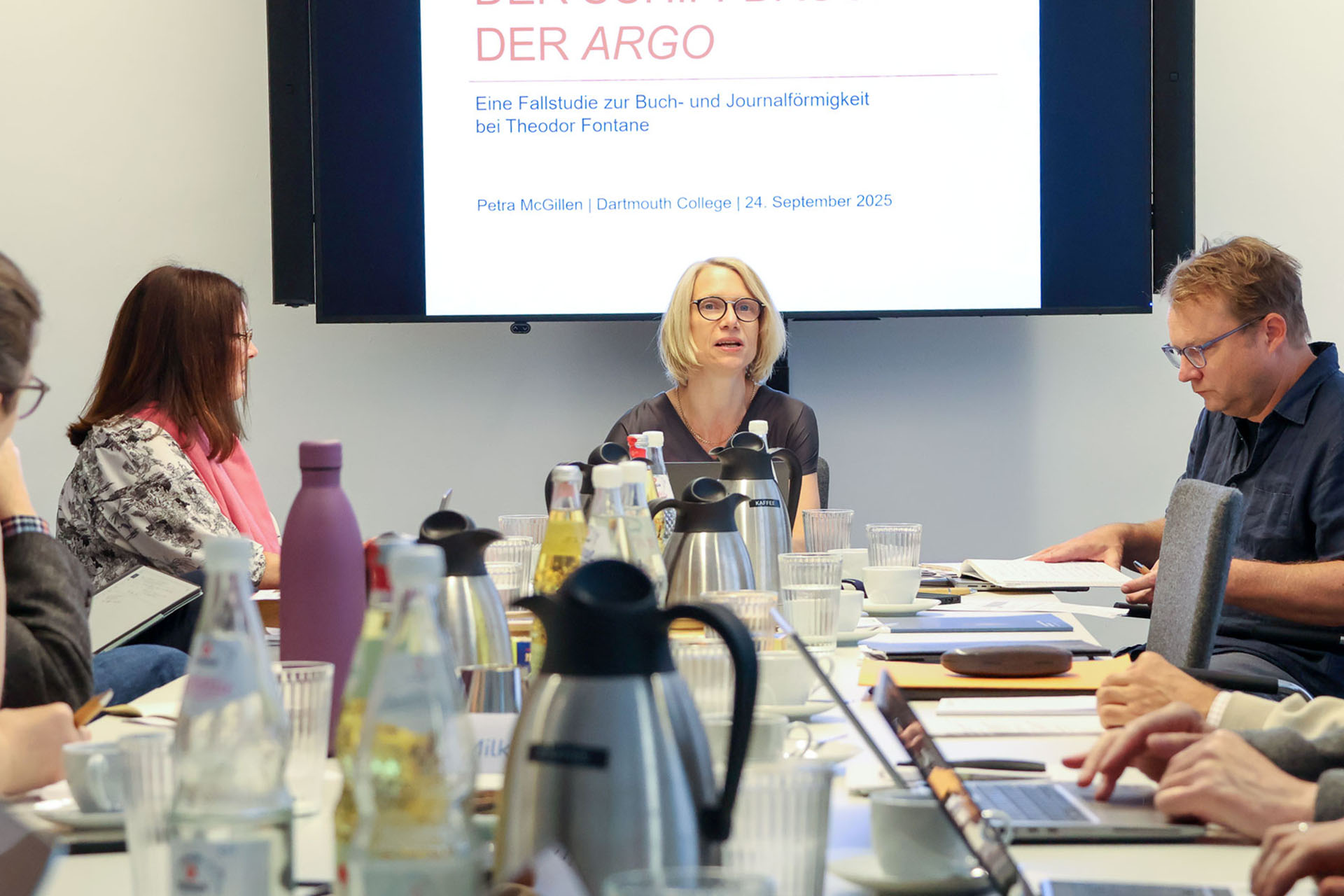 photo from the workshop 'Editing Journal Literature', participants sitting around a table, a speaker presenting, a screen with slides in the back