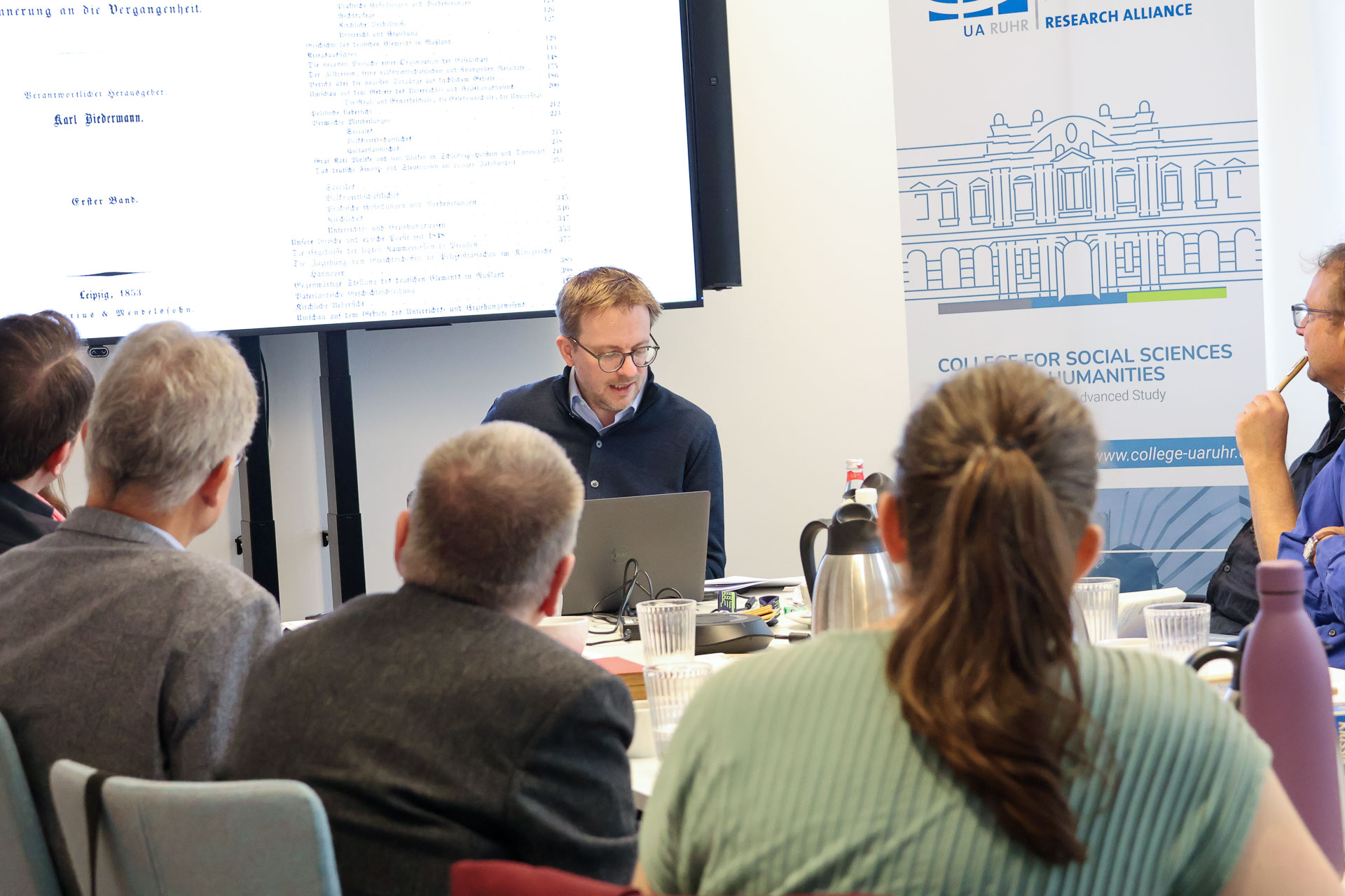 photo from the workshop 'Editing Journal Literature', participants sitting around a table, a speaker presenting, a screen with slides in the back