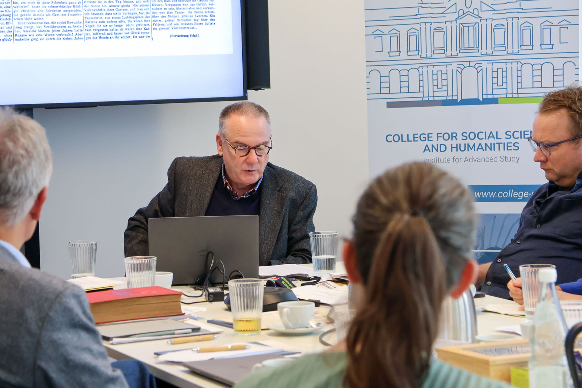 photo from the workshop 'Editing Journal Literature', participants sitting around a table, a speaker presenting, a screen with slides in the back
