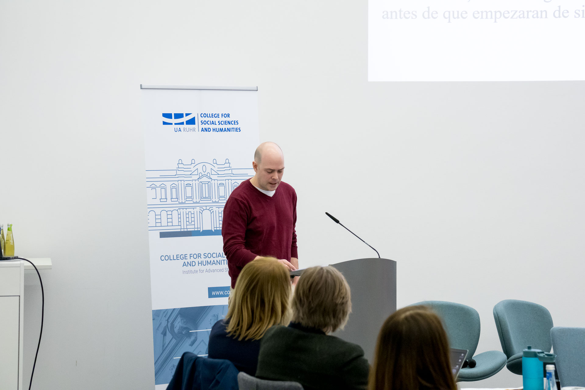 photo from the workshop 'Perpetrator Memory in Spanish culture', participants sitting around a table, a speaker presenting, a screen with slides in the back