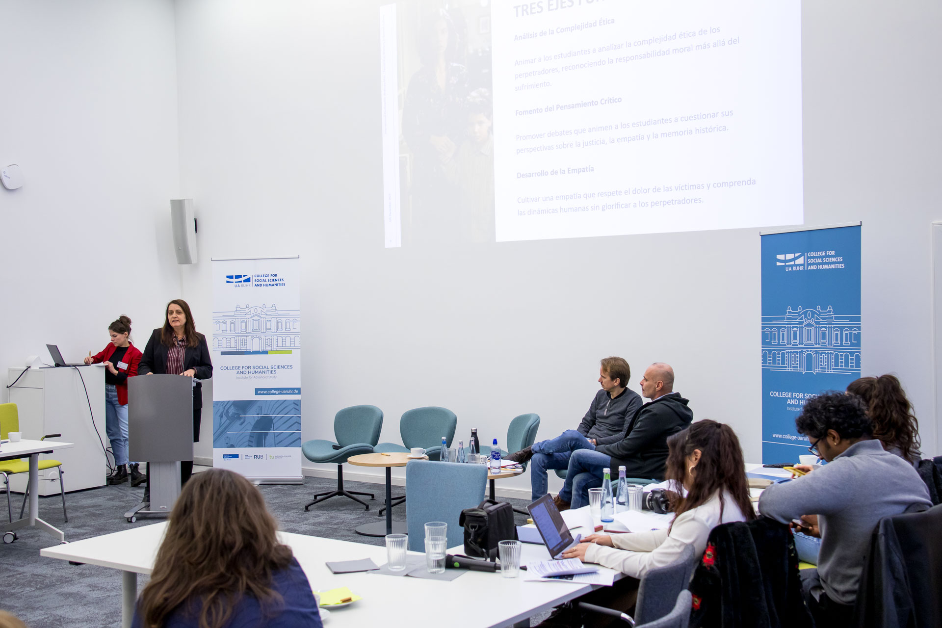 photo from the workshop 'Perpetrator Memory in Spanish culture', participants sitting around a table, a speaker presenting, a screen with slides in the back