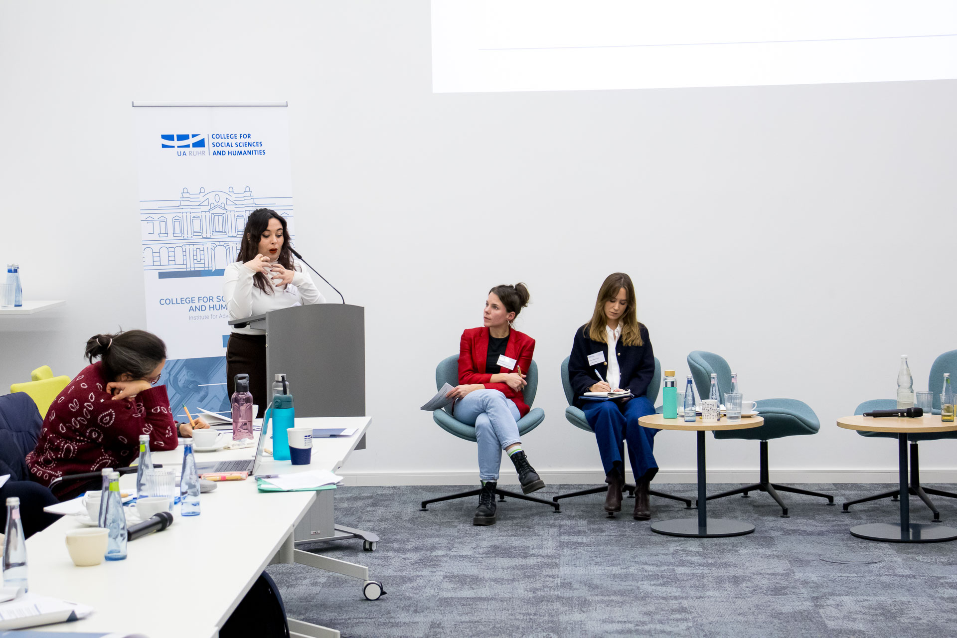 photo from the workshop 'Perpetrator Memory in Spanish culture', participants sitting at a table, a speaker presenting, a screen with slides in the back