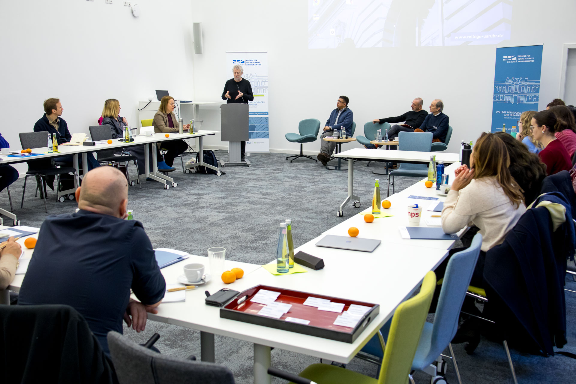 photo from the workshop 'Perpetrator Memory in Spanish culture', participants sitting at a table, a speaker presenting, a screen with slides in the back