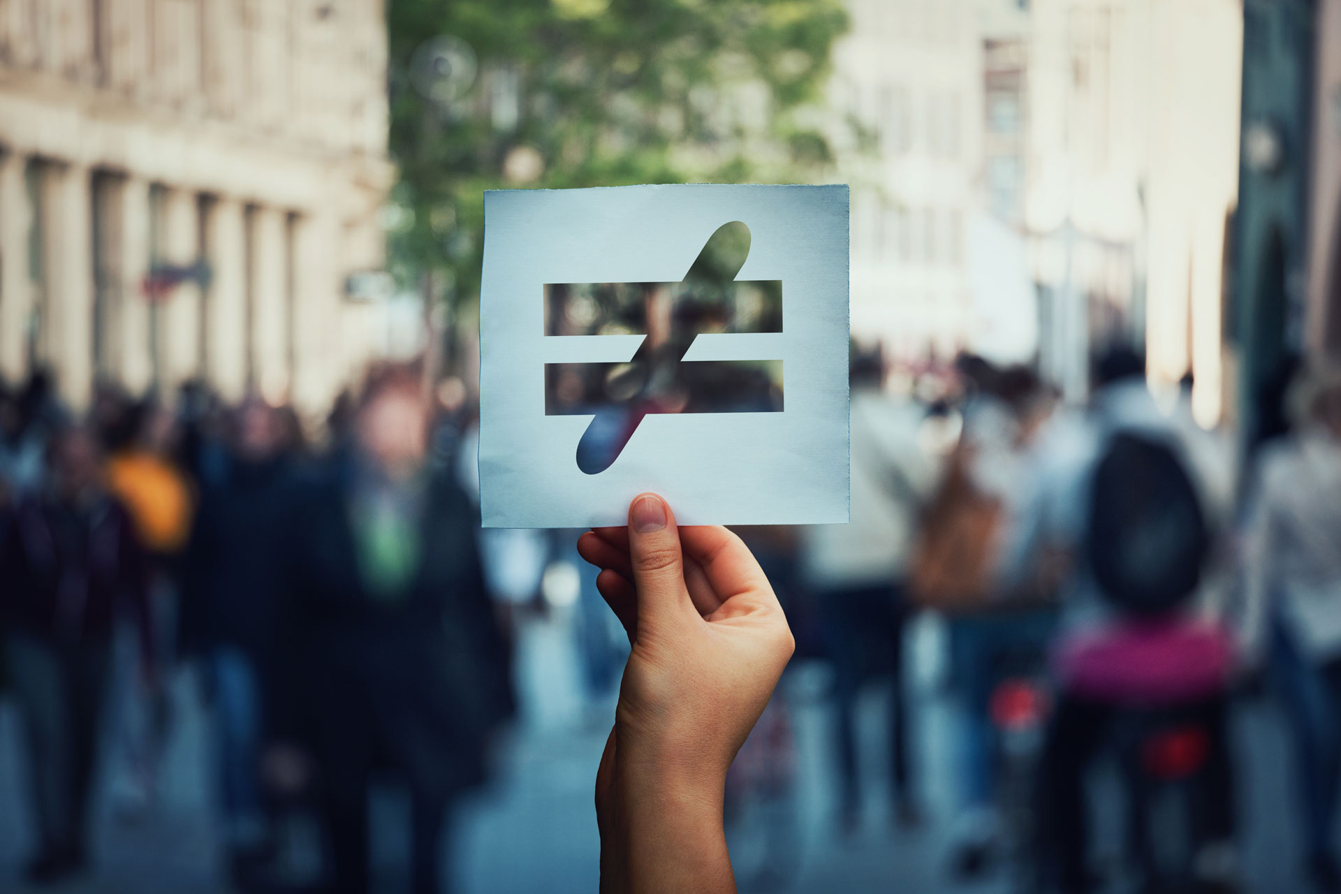 a hand holding a stenciled paper with a crossed equality symbol; blurred in the background is a crowd of people